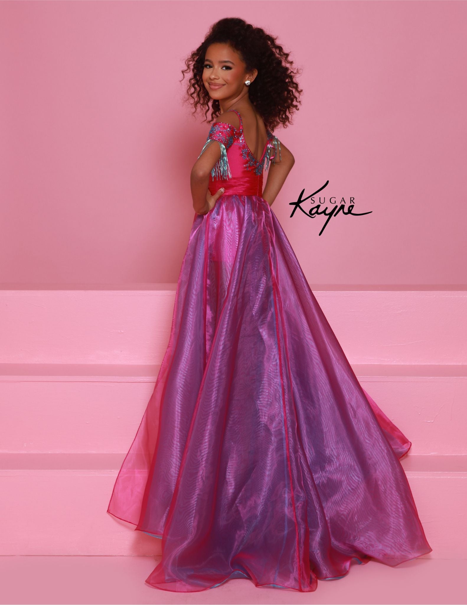 The Sugar Kayne C372 Girls Fringe Romper Overskirt offers a fun and fashionable twist on traditional pageant wear. The ombre design adds a trendy touch, while the fringes create movement and excitement on stage. Perfect for making a statement and standing out from the competition. Step onto the stage with our mesmerizing fun fashion! The romper features off-the-shoulder straps, intricate beading, and overskirt to ensure you rock the stage at your next event.

Sizes: 2-16

Colors: Magenta/Aqua